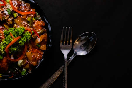 Cropped shot of tasty Chilli chicken along with fork and spoon kept on dark surfaceの写真素材