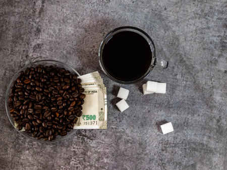 Over the top shot of black coffee, sugar cubes, and money covered with roasted coffee beans.の写真素材