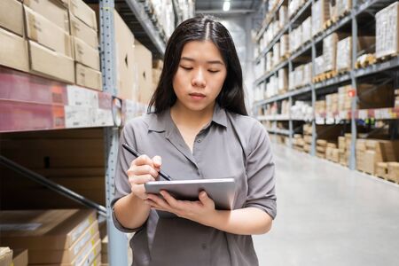 Beautiful young Asian woman auditor or trainee staff work stocktaking with computer tablet at warehouse store, Asian working business woman conceptの写真素材
