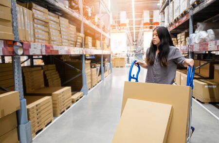 Beautiful young Asian woman customer or staff worker pushing trolley or picking cart in warehouse store with blur background of boxes on shelf, self service shopping conceptの写真素材