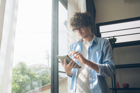 Businessman or creative freelancer man in eyeglasses standing at the window using digital tablet working for customer order or browsing information on the internet at home.の写真素材
