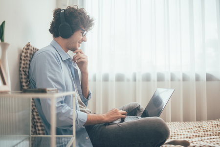 Happy freelancer man listening to music with headphone sitting on bed working with laptop or video conference meeting with customer. Communicating in social network, searching information onlineの写真素材