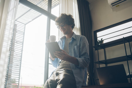 Businessman or creative freelancer man in eyeglasses drinking coffee sitting on the desk using digital tablet working for customer order, reading, browsing information on the internet at home.の写真素材
