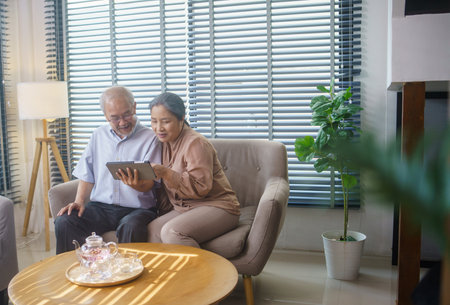 Asian grandfather and grandmother watching or meeting video call to family with digital tablet in the living room. Senior couple playing social media or searching on the internet.の写真素材