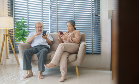 Grandfather and grandmother using smartphone watching video clip or reading message sitting on couch in the living room. Senior couple connecting with social media and technologyの写真素材