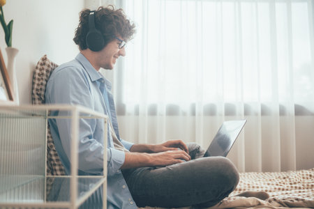 Happy freelancer man listening to music with headphone sitting on bed working with laptop or video conference meeting with customer. Communicating in social network, searching information onlineの写真素材
