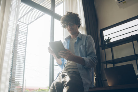 Businessman or creative freelancer man in eyeglasses drinking coffee sitting on the desk using digital tablet working for customer order, reading, browsing information on the internet at home.の写真素材
