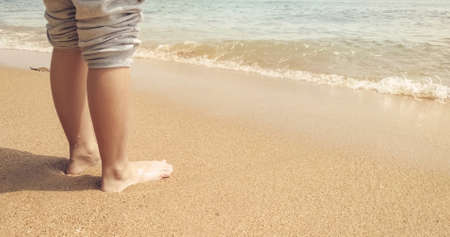 A child's wet feet relaxing on the sand. Kid on vacation. Sea waves on the tropical beach. Warm natural sunlight and clear water.の写真素材