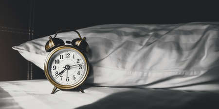 The morning sun shines in the dark bedroom. The classic alarm clock and pillow on the bed. Time to...