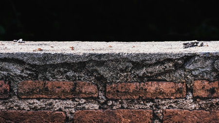 Aged red brick wall with a rough cement top, isolated against a dark background. Foreground obstacle barrier. Architectural and vintage backdrop.の写真素材