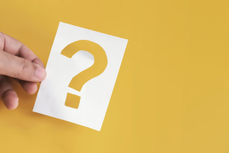 Hand holding paper cut question mark on yellow background. Concept of education, asking questions, problem solving, FAQ, consulting and seeking adviceの写真素材