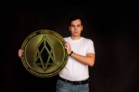 A young guy holds a symbol of the EOS coin, a modern currency for exchange and purchases. Photo on a...