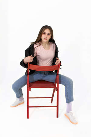 A girl in jeans sits on a chair and straightens her jacket. Photo on a white backgroundの写真素材