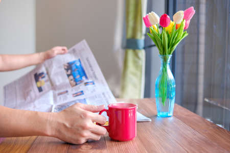 Someone picks a hot coffee while he reads newspaper.
