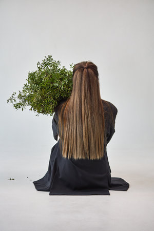 A woman with long straight hair sits facing away, holding a lush bouquet of greenery, against a plain studio background.の写真素材