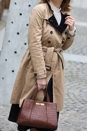 Stylish woman wearing beige trench coat, holding brown leather bag, standing outdoors on sunny day, elegance, fashion, style, autumn wardrobe conceptの写真素材