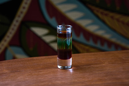 A visually striking layered cocktail served in a shot glass, placed on a rustic wooden table. Ideal...