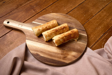 Three crispy, golden-brown fried spring rolls placed on a rustic wooden cutting board, set against a...
