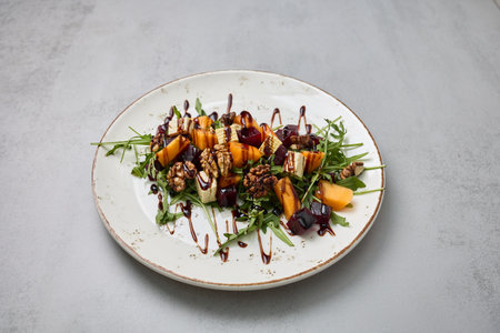 A fresh salad with arugula, persimmon, beetroot, walnuts, and feta cheese, drizzled with balsamic...