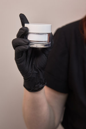 Hand in black glove holding white cosmetic jar, closeup of beauty product in professional care setting, skincare focus conceptの写真素材