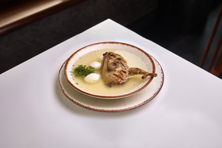 A bowl of clear quail broth featuring grilled quail leg, quail eggs, and fresh herbs served on a rustic ceramic dish.の写真素材