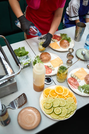 Restaurant staff in gloves pours soda into citrus mint drinks while prepping burger buns, cheese...