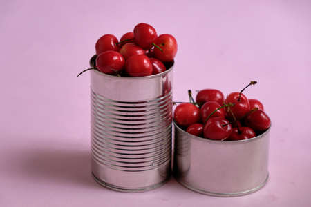A closeup shot of two metal cans filled with fresh cherries on a pink surface