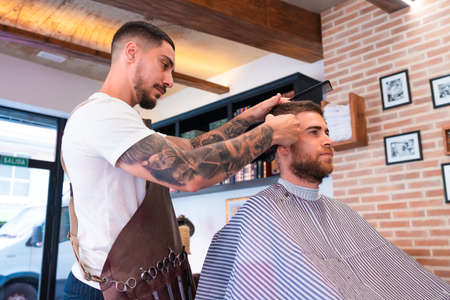 A young hairdresser cutting the male customer's hair with a hair clipper and a comb in a barbershopの写真素材