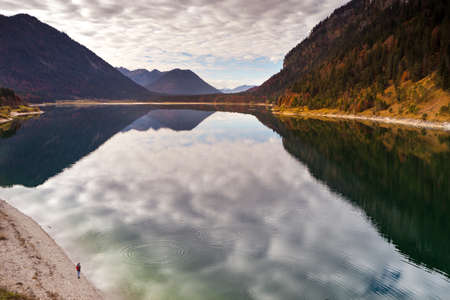 A beautiful scenery of high rocky mountains with green trees reflecting in the lake under a cloudy skyの写真素材