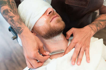 A high angle view of a hairdresser with tattoos shaving the beard of a customer with a bladeの写真素材