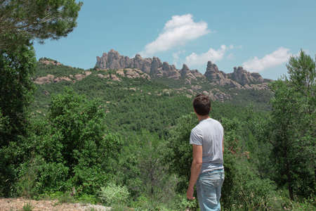 A side view of a young male wearing a gray shirt and jeans shorts posing in front of scenic green...