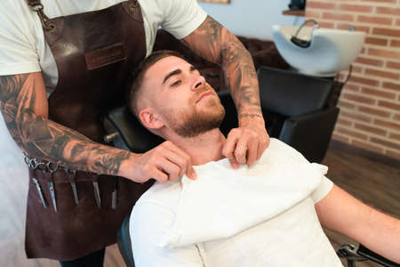 A high angle view of a young hairdresser with tattoos putting a towel on the male customer's chest before shavingの写真素材