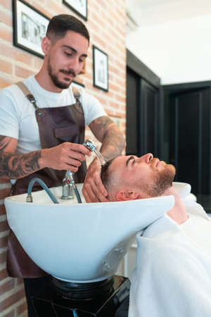 A vertical shot of a hairdresser with tattoos washing the hair of a young male customer