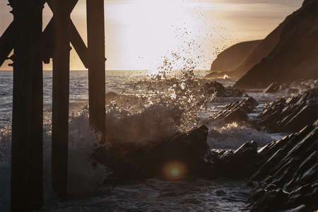 The beautiful ocean waves on spiky rocks at the coast of the sea under the wooden pier during sunset