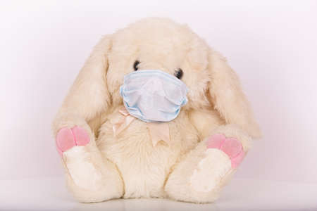 A fluffy stuffed bunny toy wearing a protective medical face mask on a white background - global...