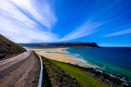 A dirt road near a grassy hill and the sea with a mountain in the distance under a blue skyの写真素材