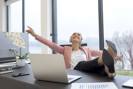 A businesswoman listening to music happily with headphones with legs on the table during the breakの写真素材