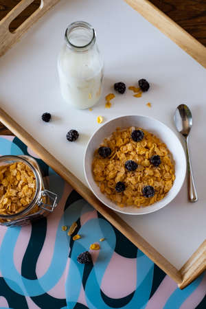 A bowl of corn flakes with blackberries next to the milk and corn flake jars