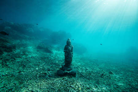 A beautiful underwater shot of an old Asian statue surrounded by fish with sunlight shining on itの写真素材