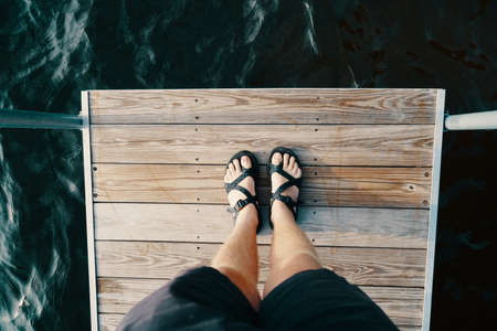 The feet of a male standing on a wooden surface over the body of waterの写真素材
