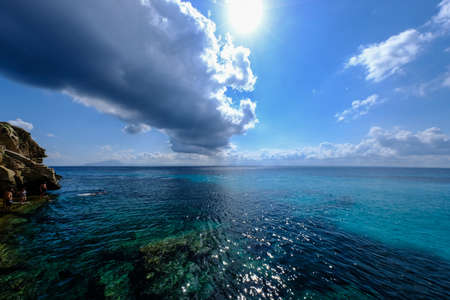 A wide shot of a clear water sea with cliff and people on the side on a sunny day