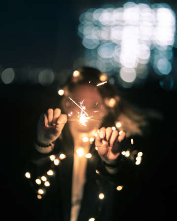Female in blur holding bright sparkle sticks in focus close to the camera at nightの写真素材