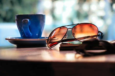 A closeup moody bokeh shot of aviator type of glasses with a blue cup of coffee with a blurred backgroundの写真素材