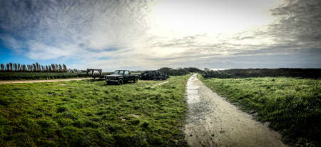 A panoramic shot of a green field with a narrow road in the middle and pickup truck parked on the...