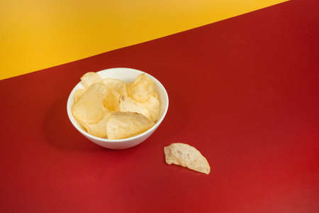 A high angle shot of plain potato chips in a white glass bowl on a yellow red background