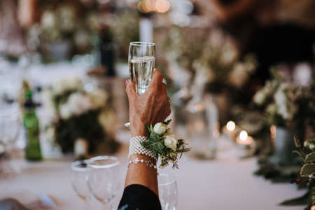 A closeup shot of an old female holding a glass of champagne with a flower pinned on an accessory on her handの写真素材
