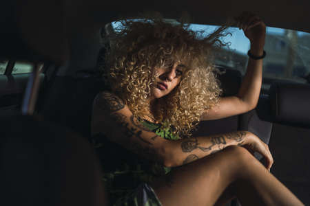 A sexy Caucasian female with curly blond hair and tattoos posing in the backseat of a car