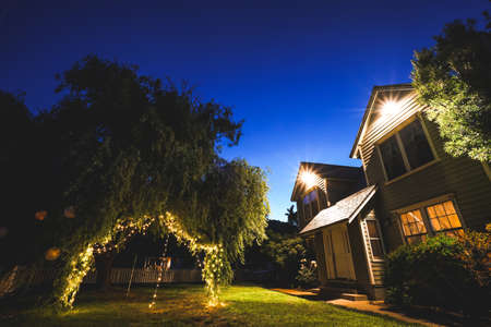 A low angle shot of a backyard with hanging tree lights at night near the house with clear blue sky...
