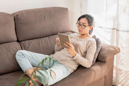 A young Asian woman lying on the couch and watching videos on a tablet