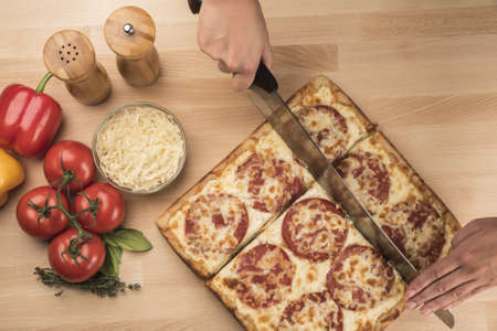 An overhead shot of a person cutting a square-shaped pepperoni pizza with vegetables on the side